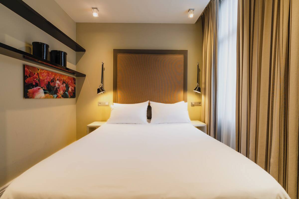 A hotel room with a large bed, a headboard with a textured pattern, bedside lamps, a shelf with decorative items, and a painting on the wall.