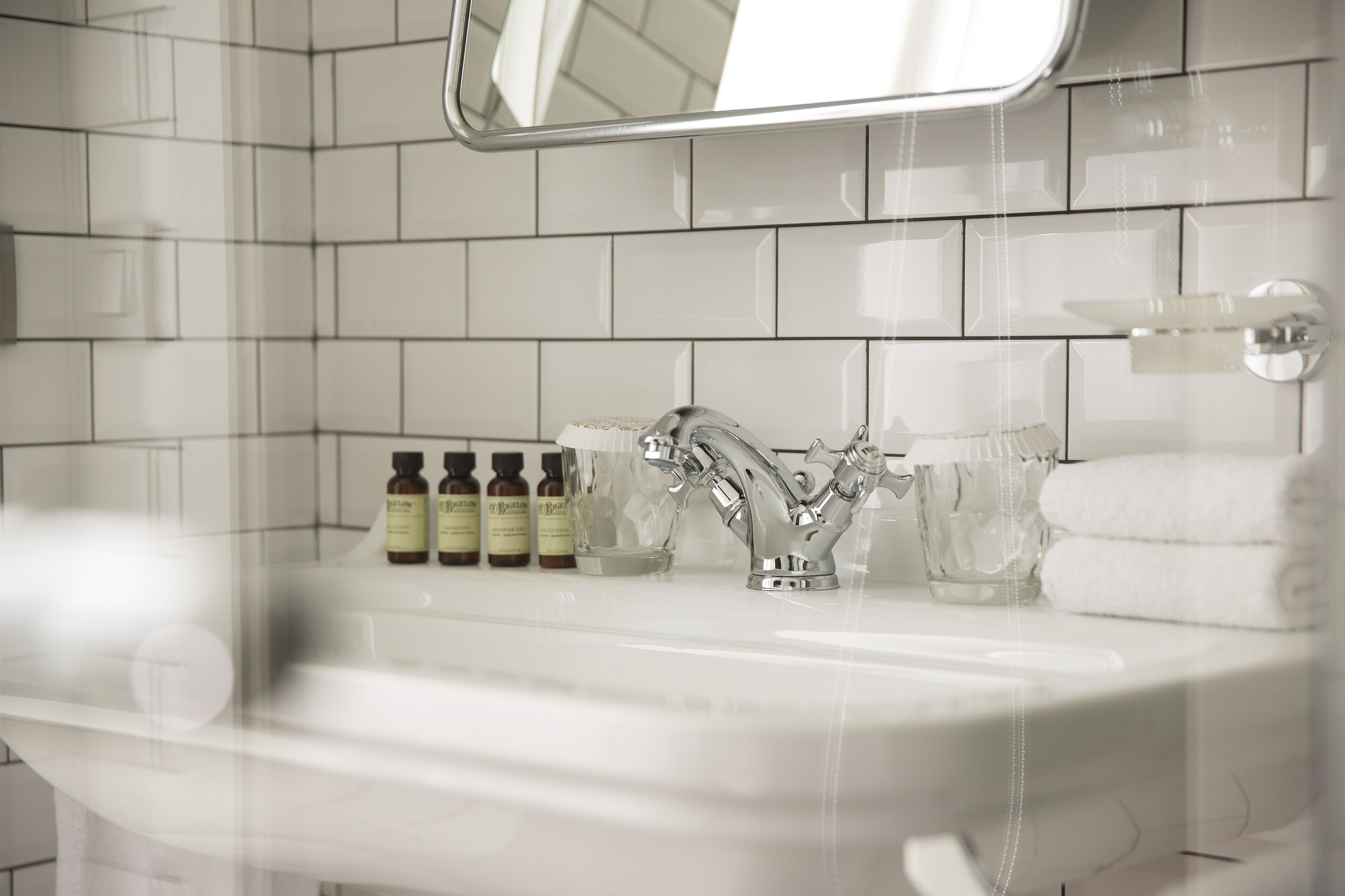 A bathroom with a white tiled wall, a chrome faucet, and a mirror above the sink. There are three bottles of lotion on the sink counter.