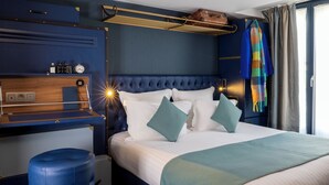 Quarto de hotel com uma cama grande, uma escrivaninha de madeira, um banquinho estofado azul e uma mala na parede.