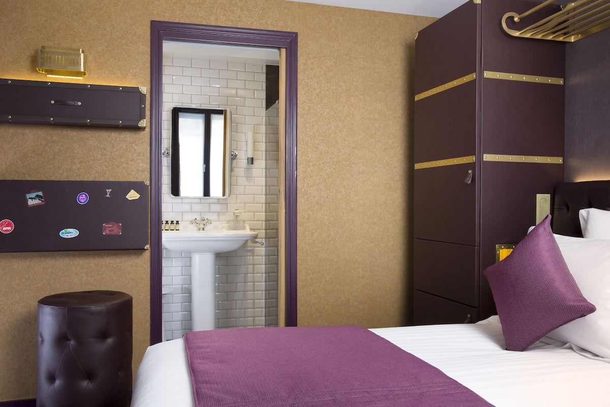 A hotel room with a bed, a purple pillow, a brown ottoman, a wall-mounted shelf with various items, and a bathroom with a sink and mirror.