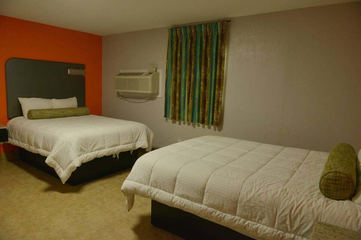 Two single beds with white bedding and green pillows, an air conditioning unit, and a window with patterned curtains.
