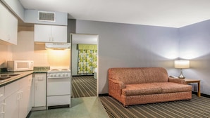 A compact hotel room with a kitchenette, a patterned sofa, and a lamp.