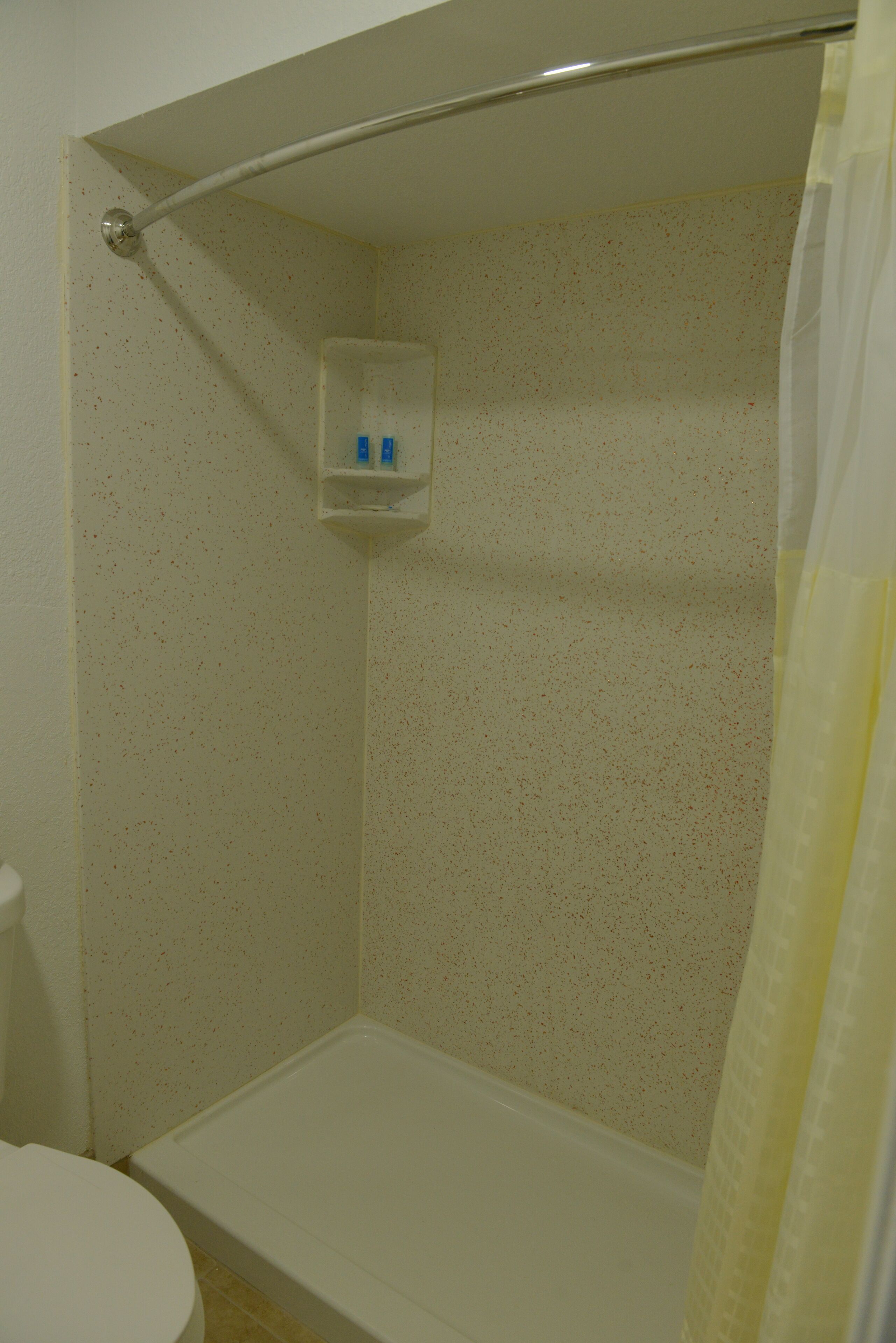 A shower with a white curtain, a small shelf with toiletries, and a plain tiled wall.