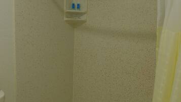 A shower with a white curtain, a small shelf with toiletries, and a plain tiled wall.