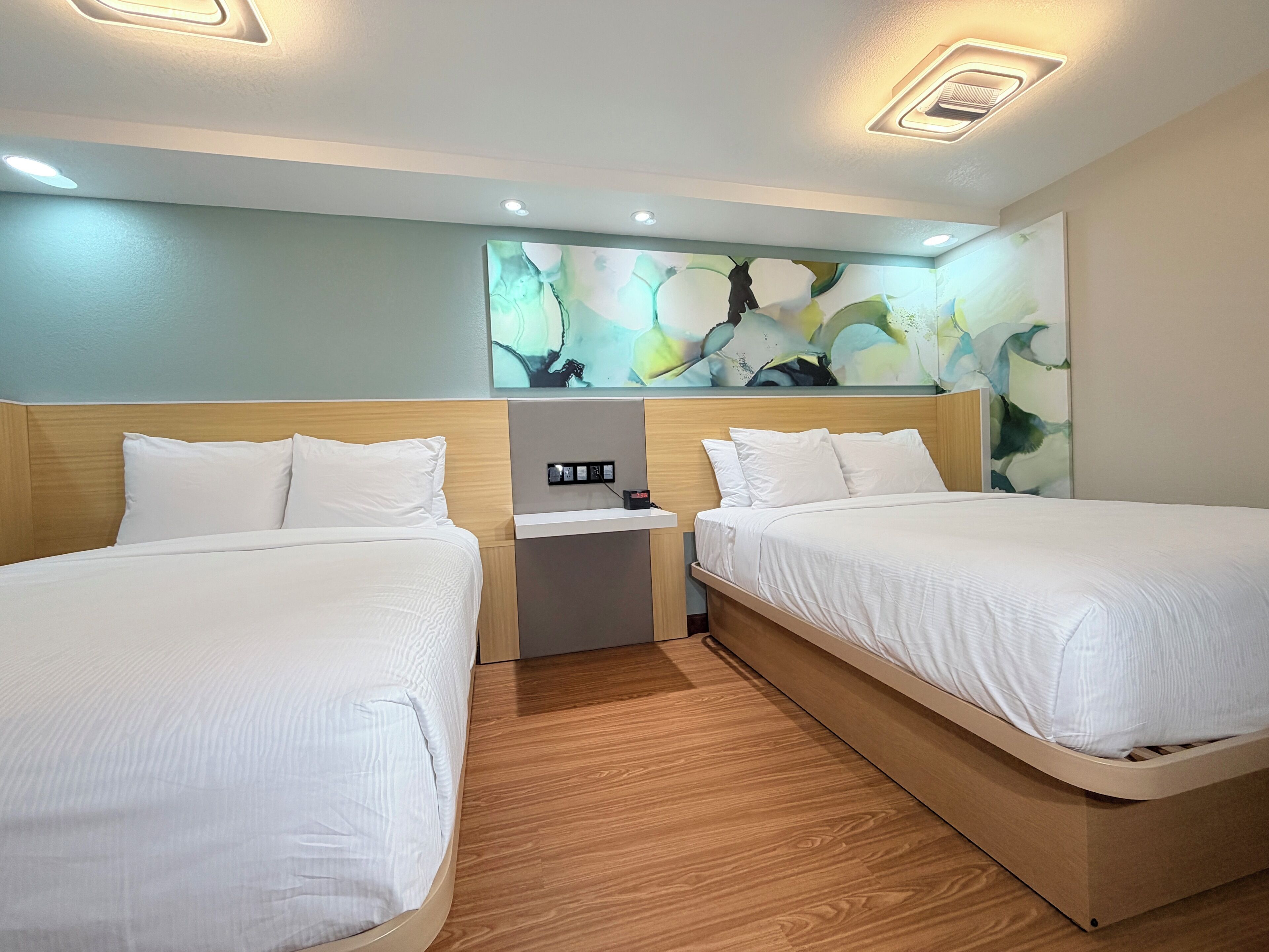Habitación de hotel con dos camas, una mesita de noche, panel de control en la pared e iluminación empotrada en el techo.