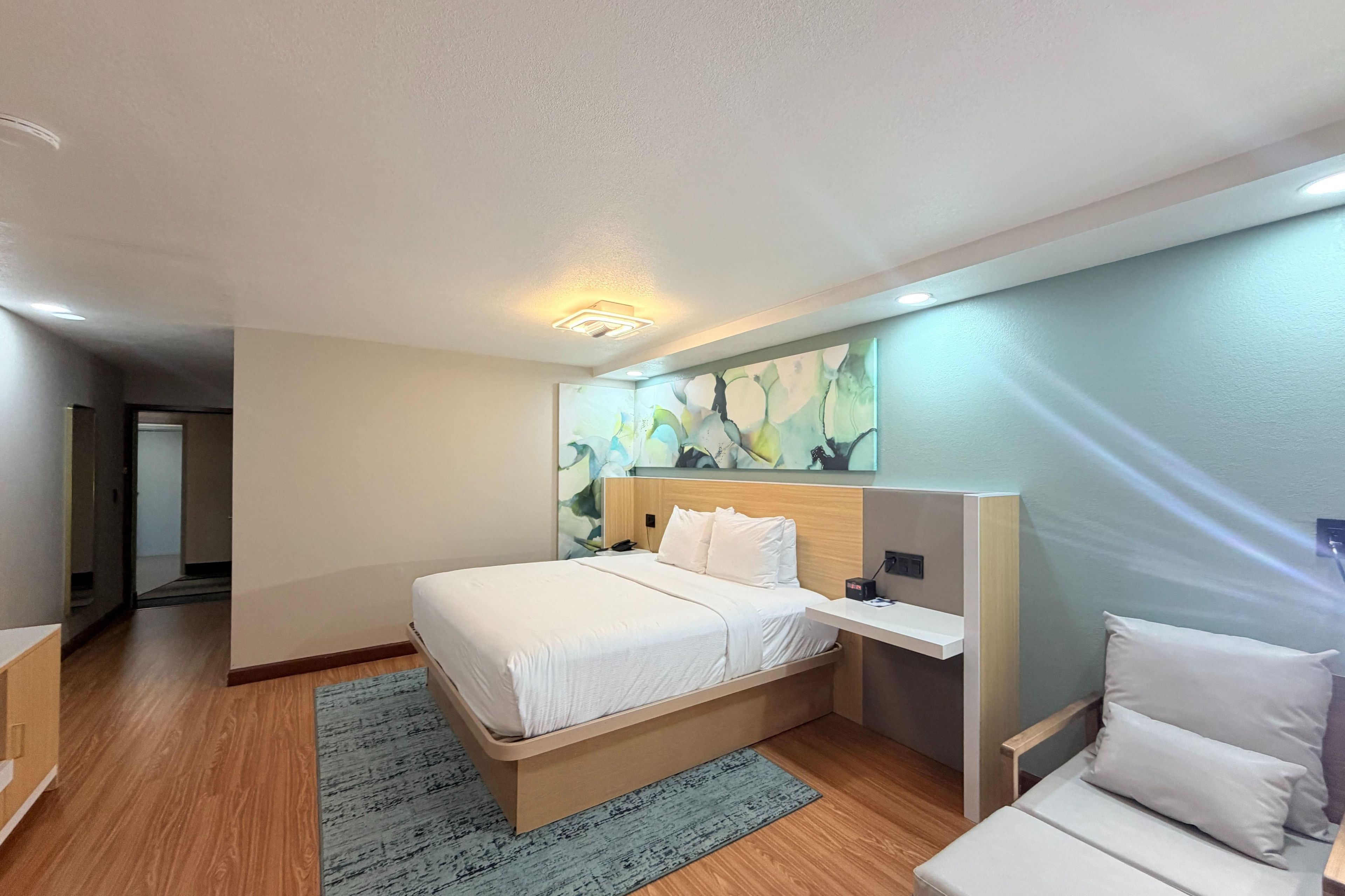 Una habitación de hotel con una cama, una mesita de noche, una obra de arte colgada en la pared y vistas al pasillo.