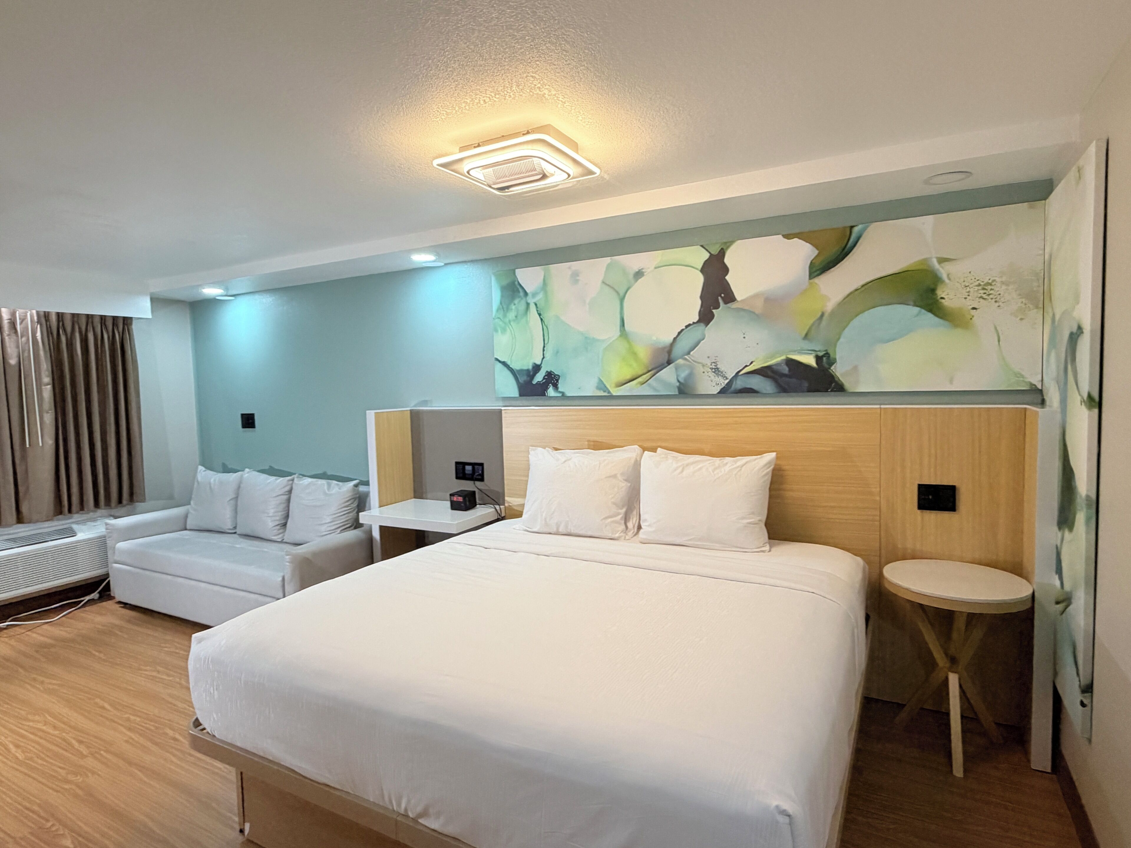 A hotel room with a large bed, a bedside table, a small round table, a sofa, and a large abstract painting on the wall.
