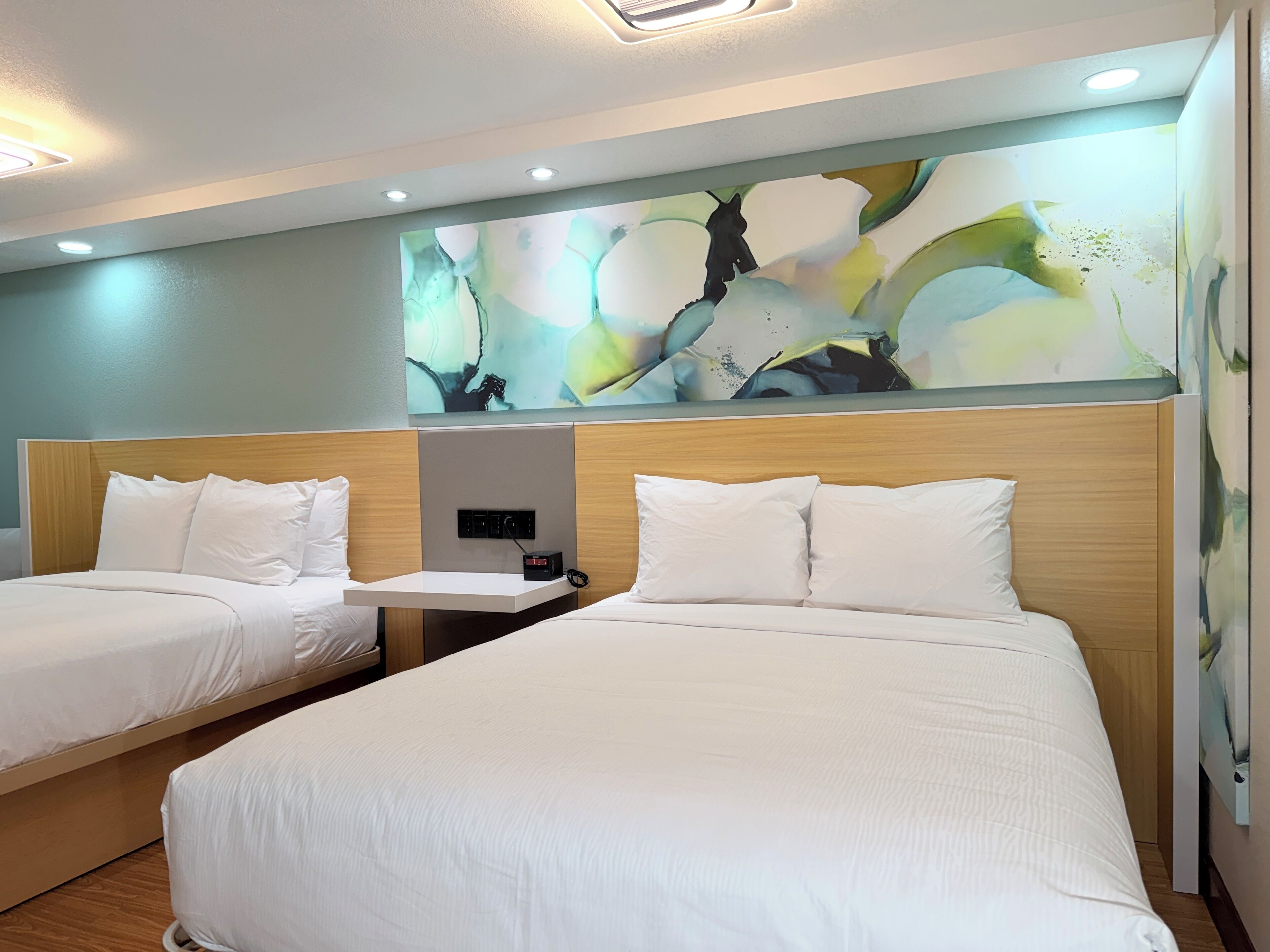 A hotel room with two beds, a nightstand, a wall-mounted control panel, and a large abstract painting.