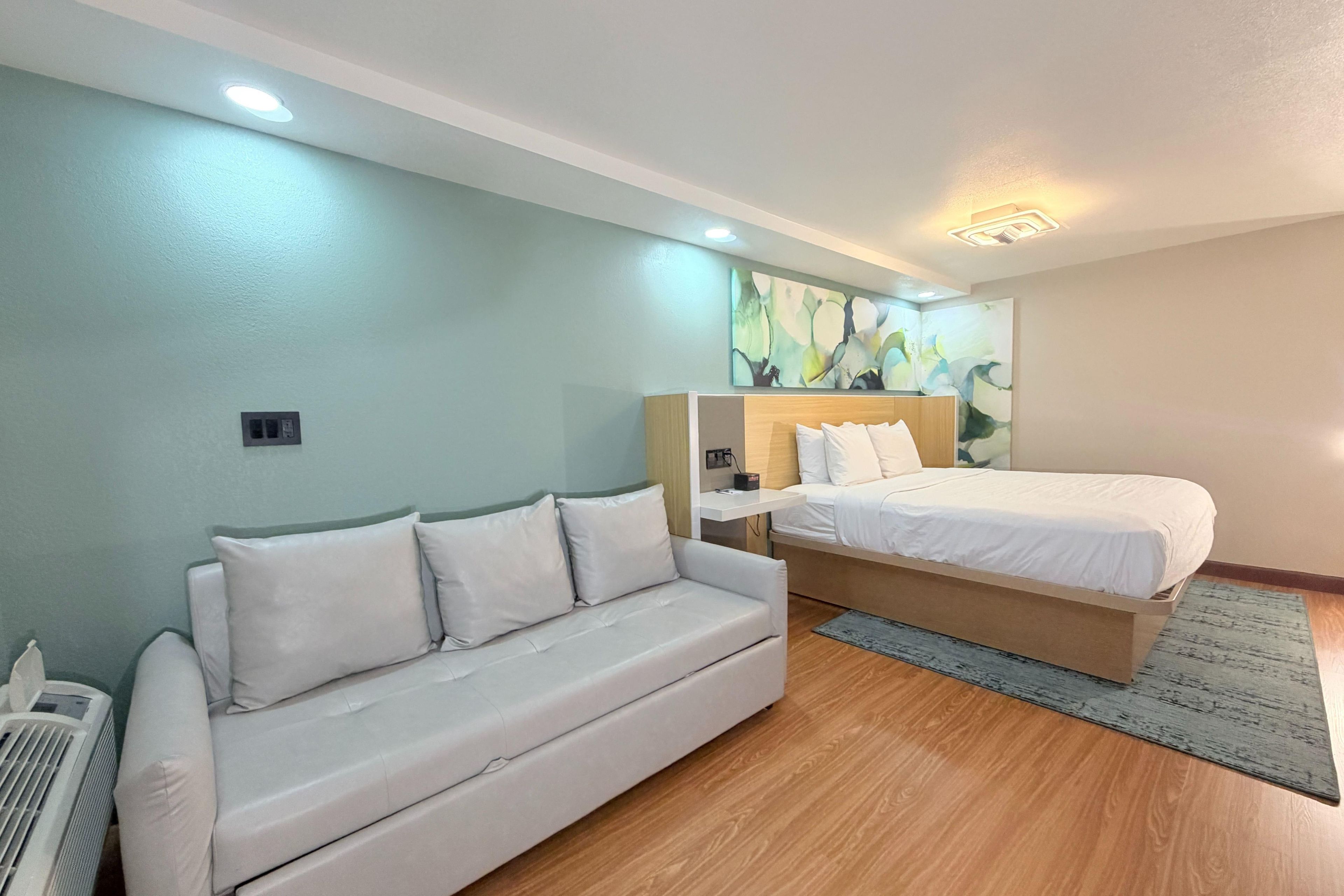 A hotel room with a bed, a sofa, a wall-mounted artwork, and a ceiling light.
