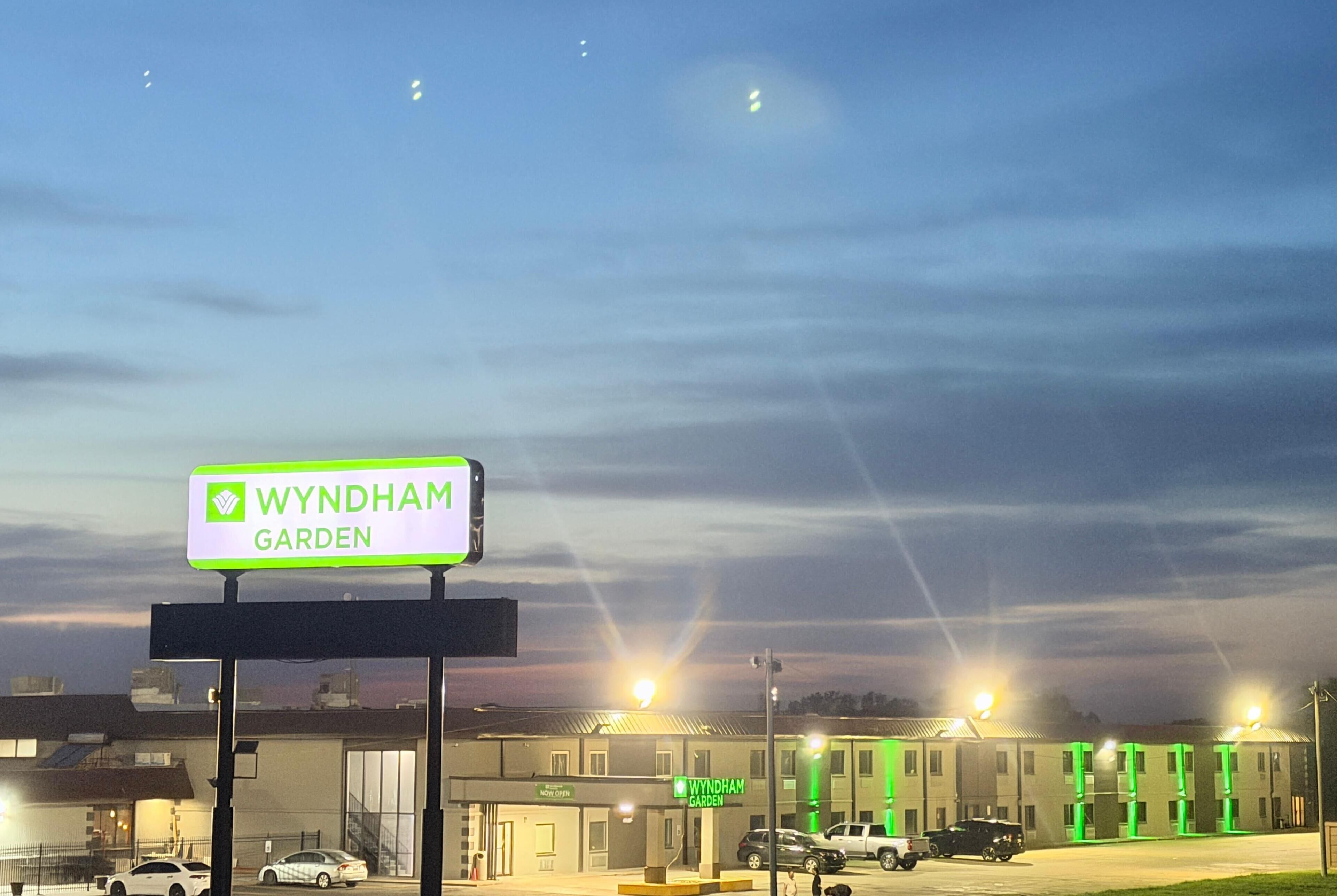 A Wyndham Garden hotel sign, a parking lot with cars, and a building with multiple windows and a green-lit entrance.