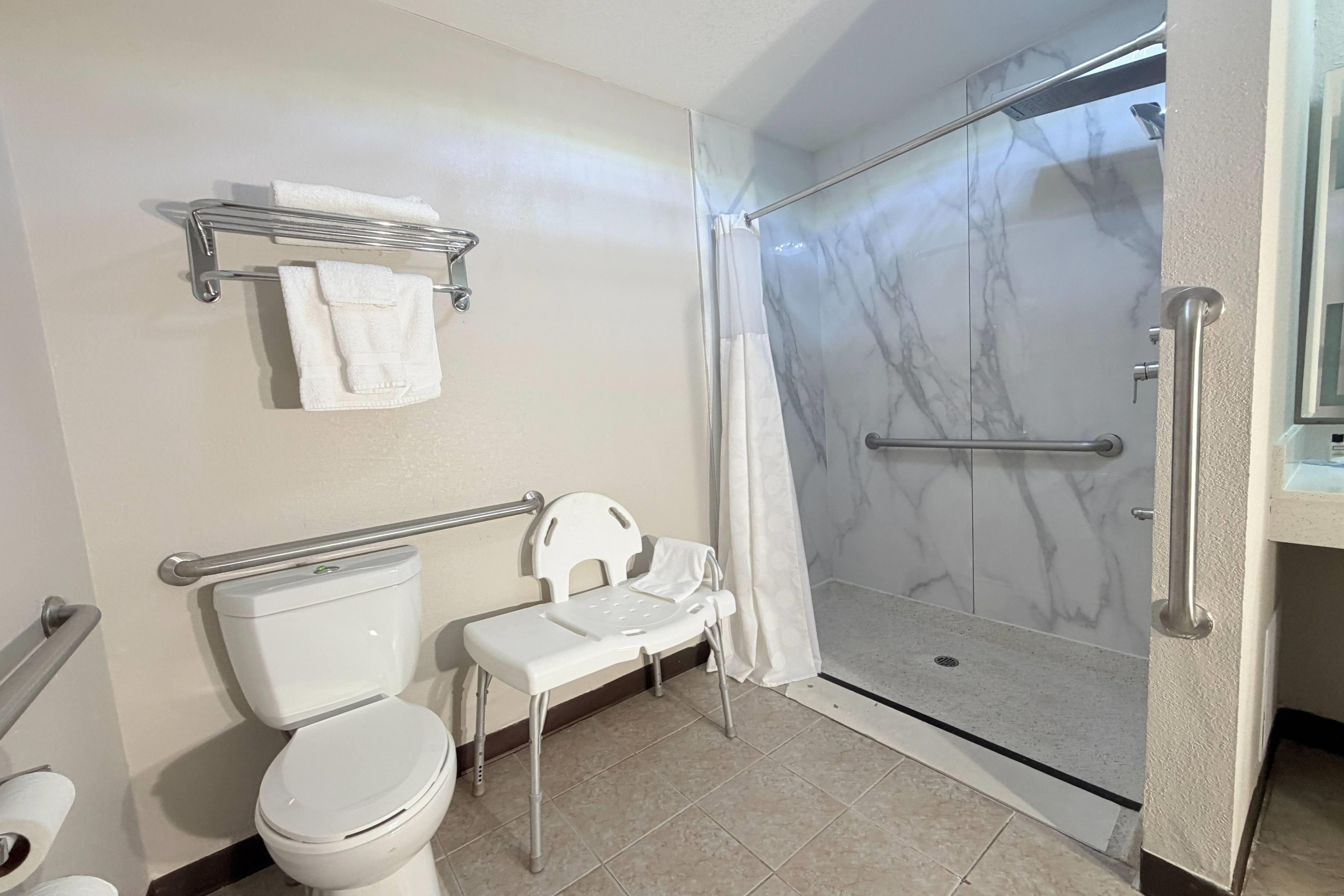 A bathroom with a toilet, a shower with a glass door, a grab bar, and a small white chair.