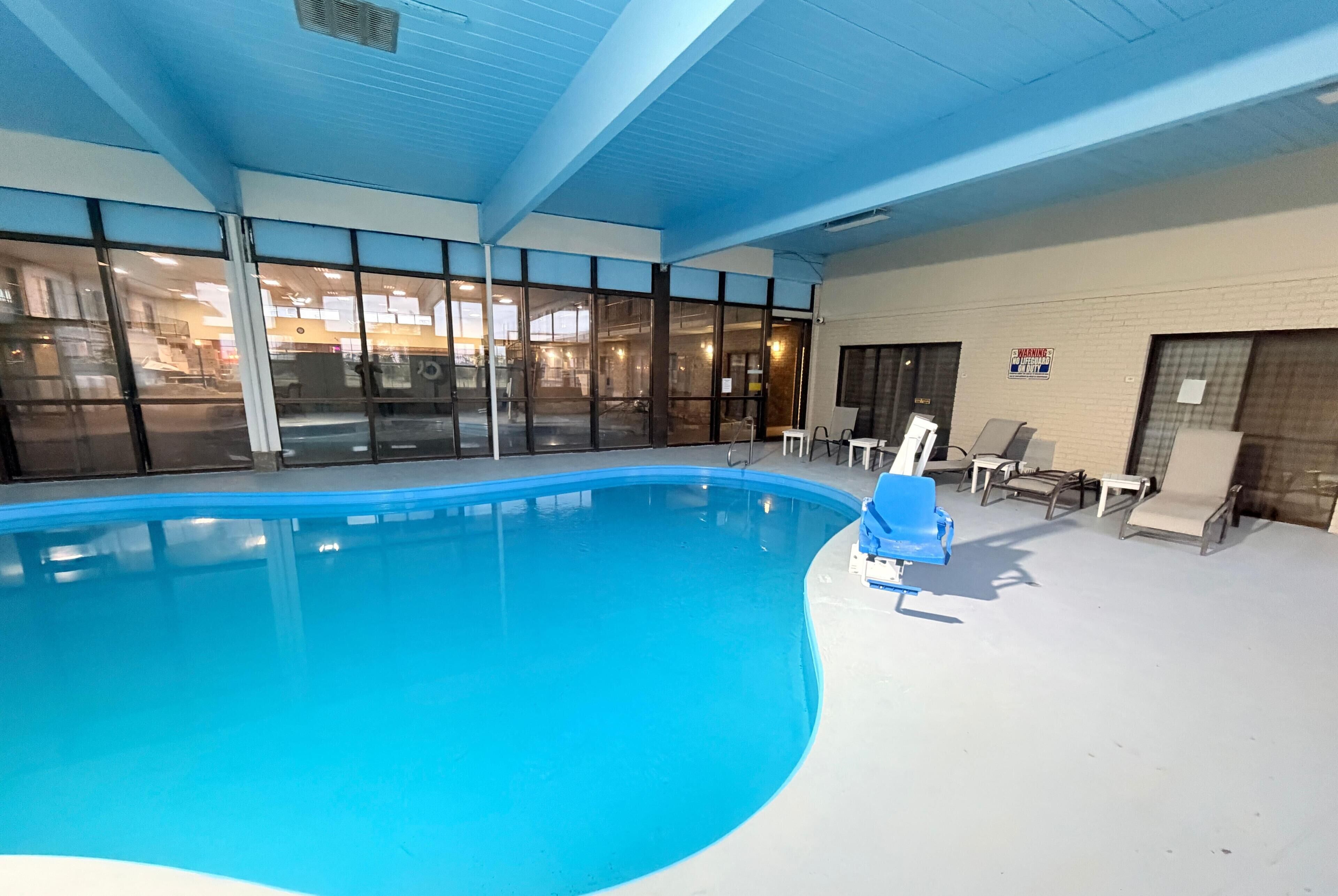 An indoor swimming pool with clear glass walls, lounge chairs, and a warning sign on the wall.