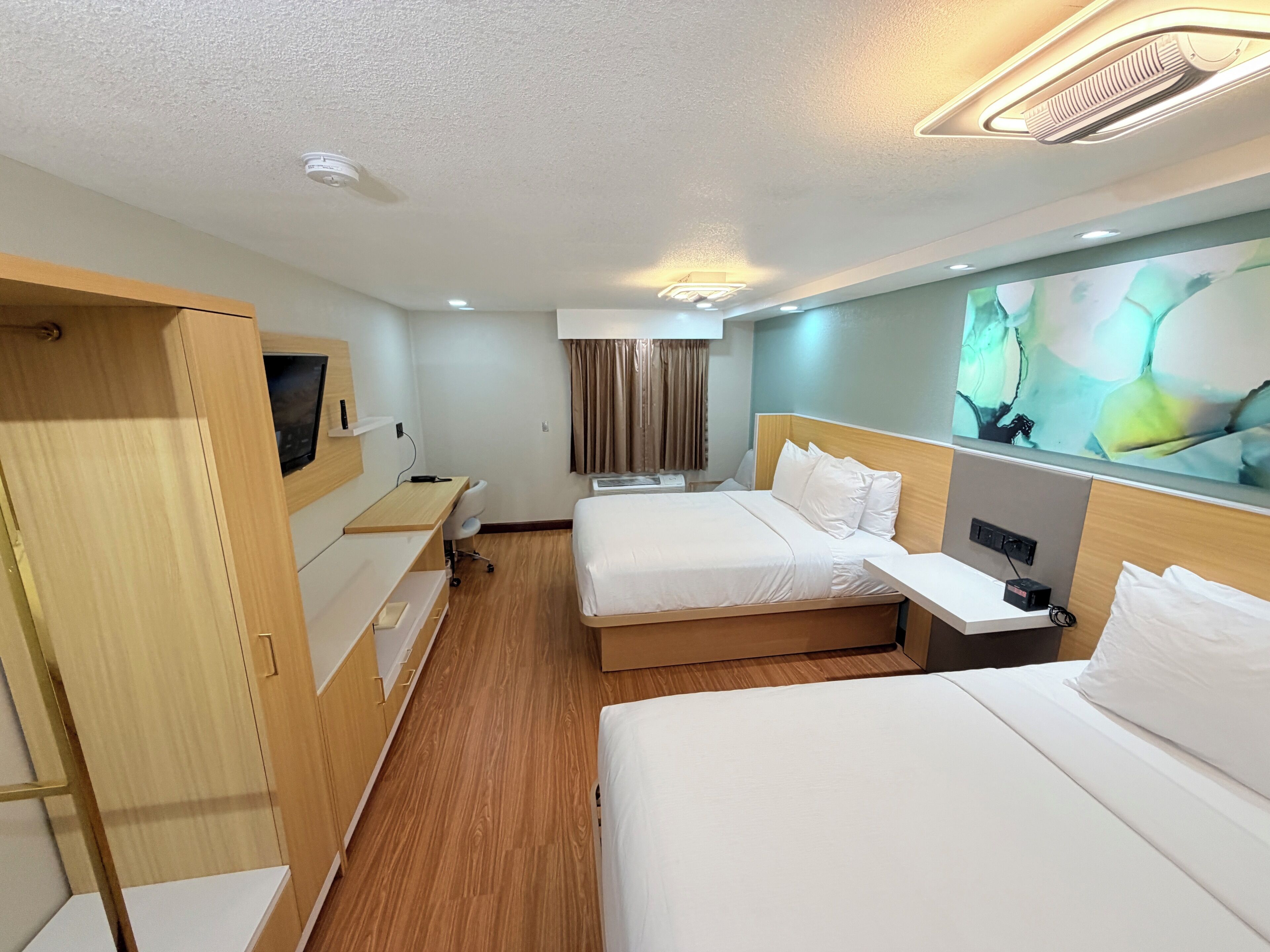 A hotel room with two beds, a desk, a wardrobe, and a large abstract painting on the wall.