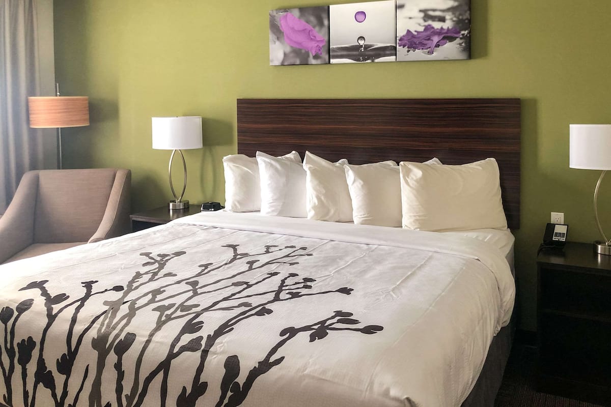 A hotel room with a large bed, a wooden headboard, two bedside tables with lamps, a chair, and a wall with artwork.