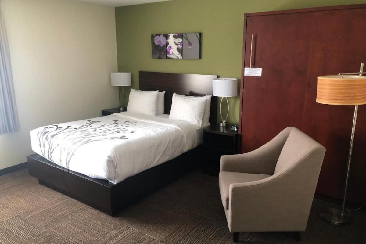 A hotel room with a bed, a chair, a nightstand, a lamp, and a door.