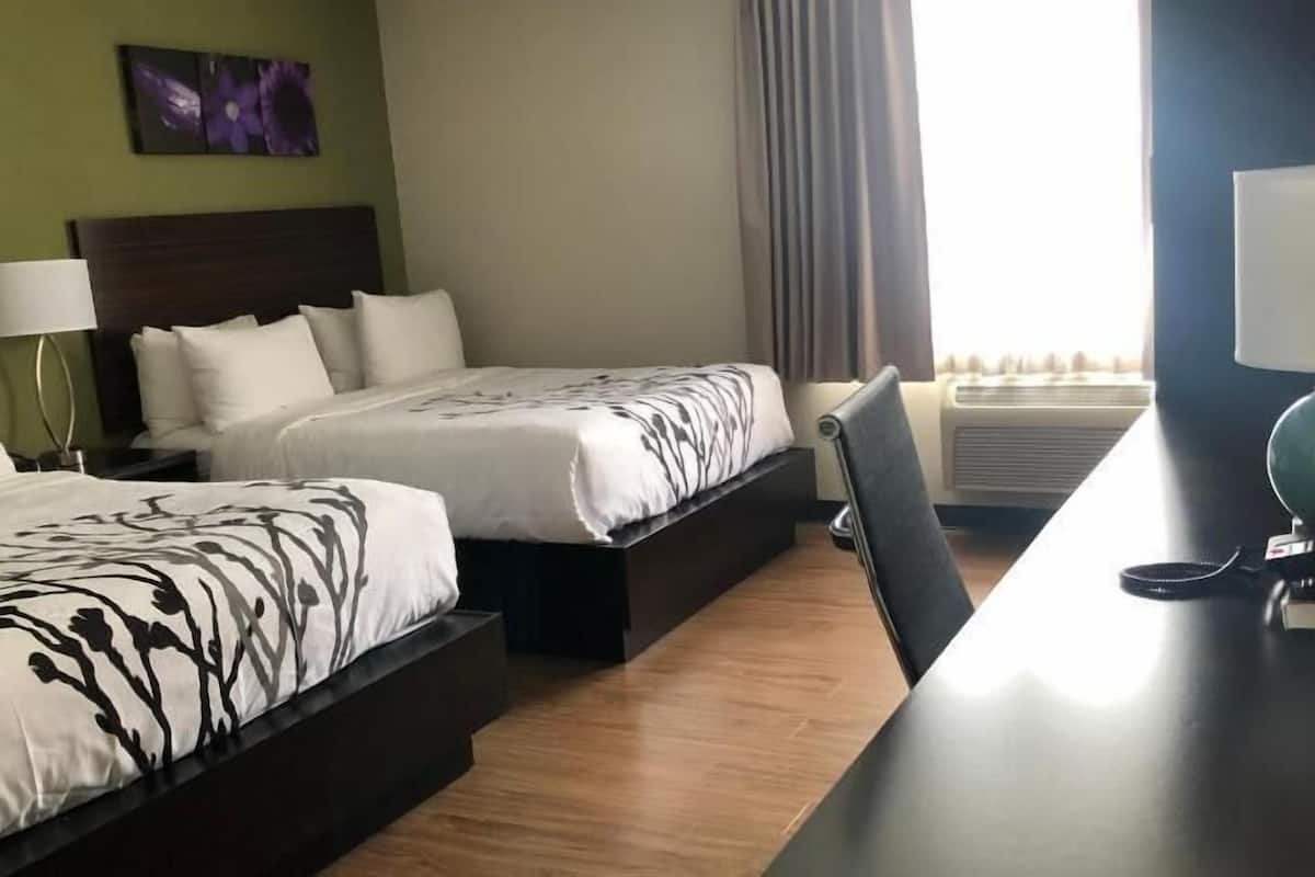 A hotel room with two beds, a desk, a chair, a lamp, and an air conditioning unit.