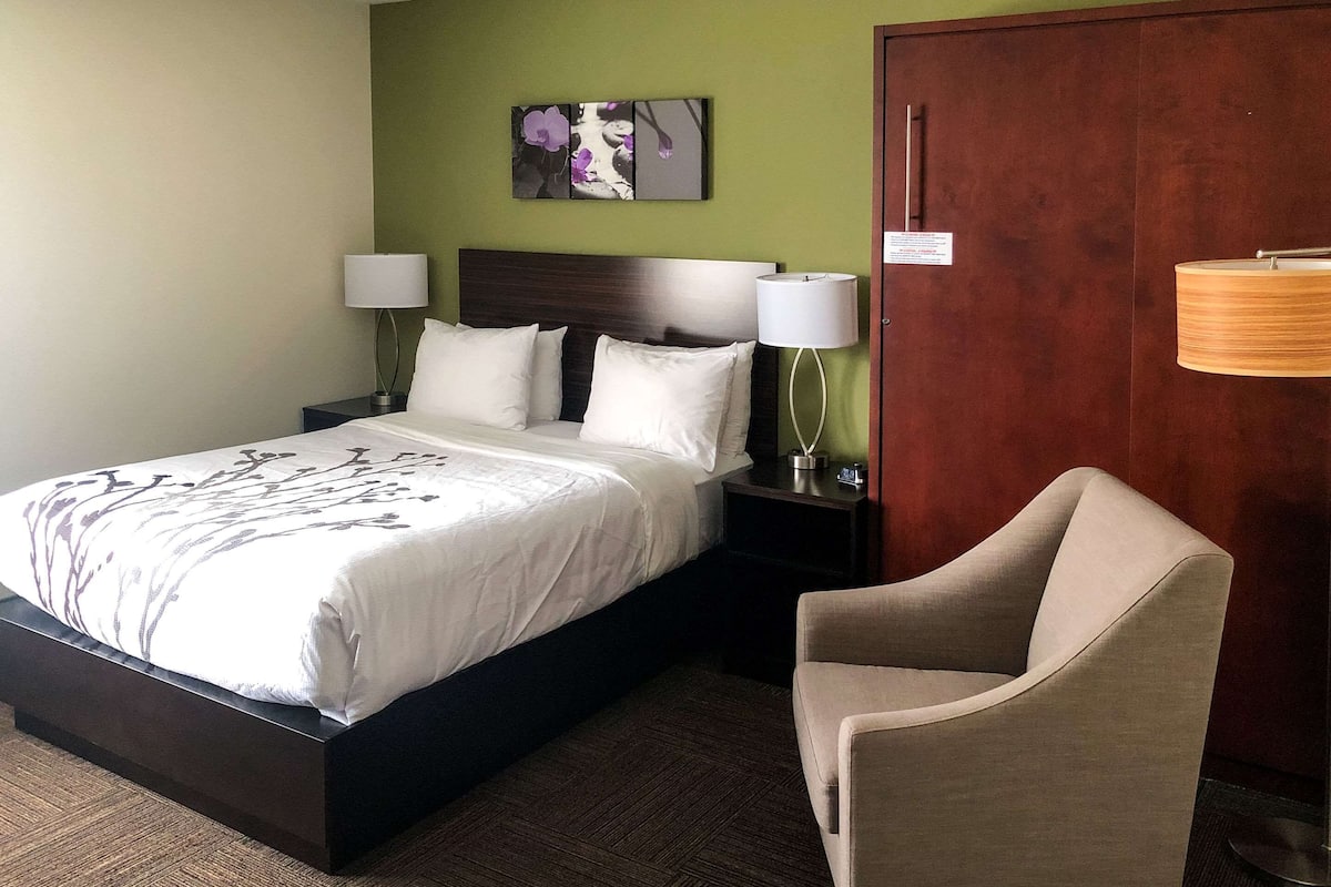 A hotel room with a bed, two bedside lamps, a chair, a wooden door, and a green wall with a floral artwork.