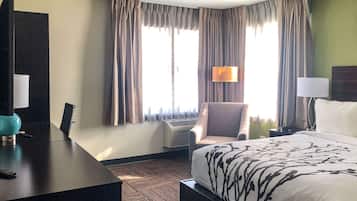 Habitación de hotel con cama, escritorio, silla, lámpara y un amplio ventanal con cortinas.