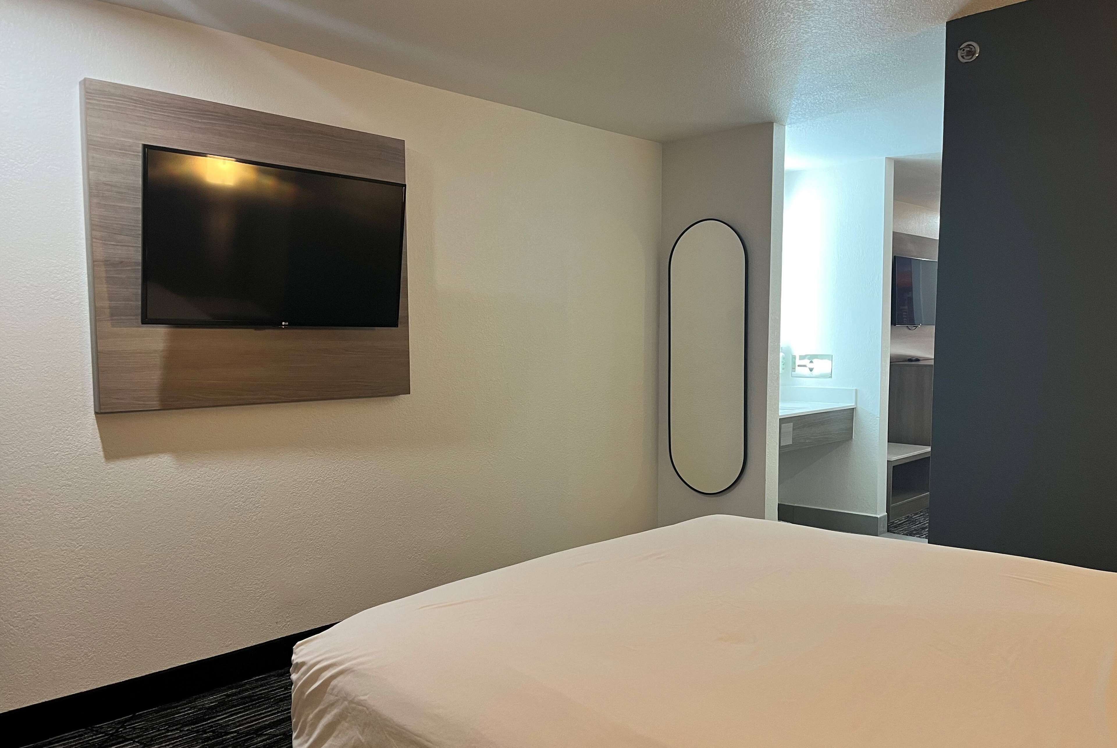 Ett hotellrum med en säng, en väggmonterad TV, en spegel och utsikt över ett badrum och en garderob.