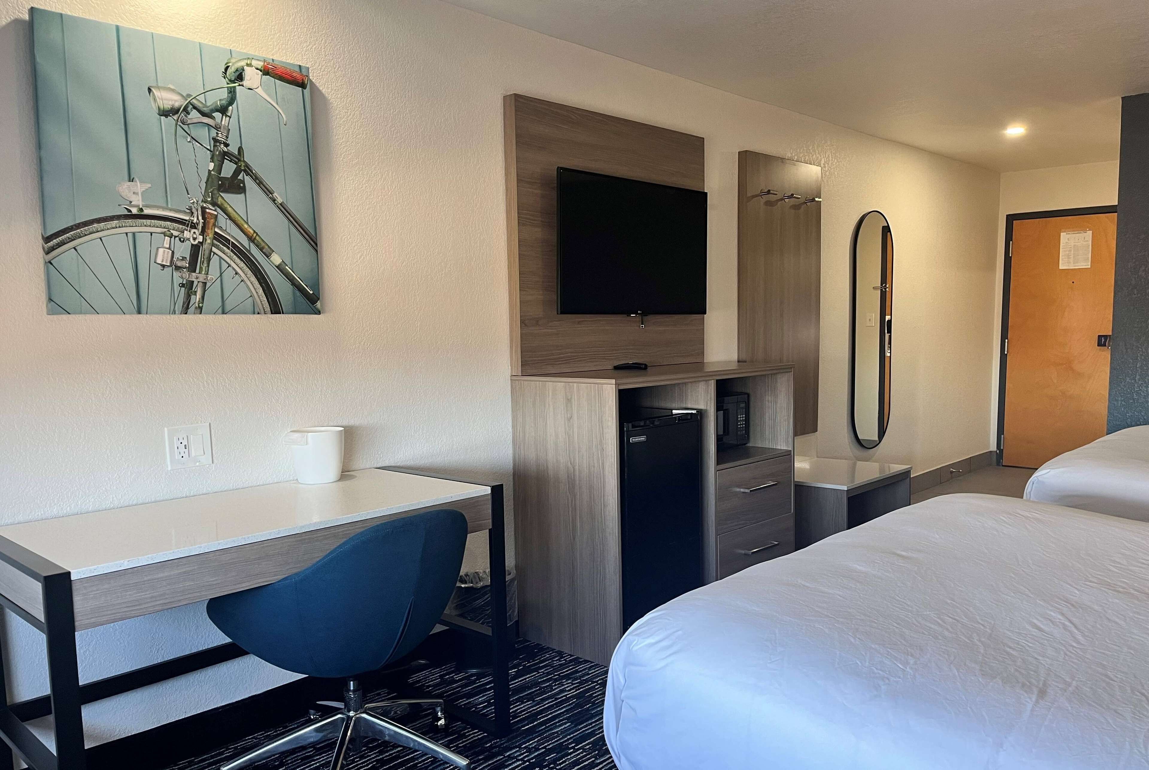 Quarto de hotel com cama, escrivaninha com cadeira, televisão, espelho e frigorífico.