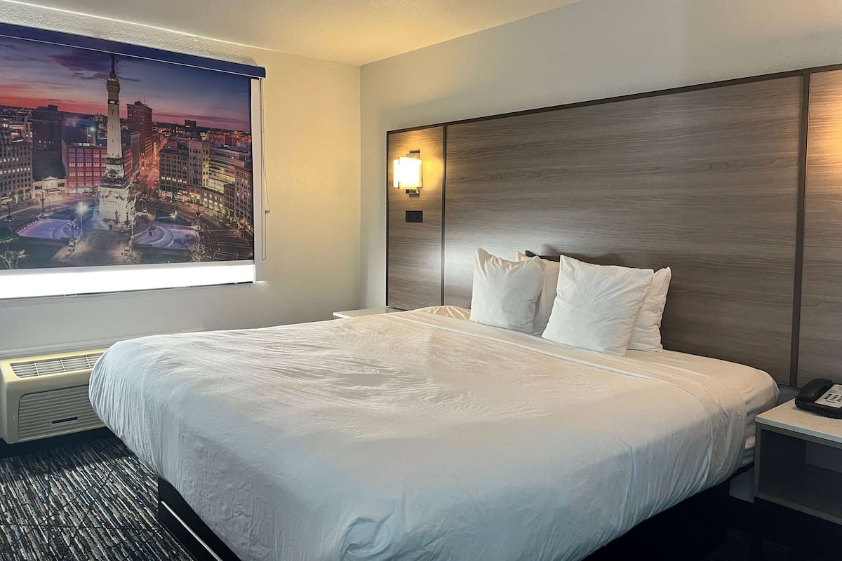 Habitación de hotel con una cama grande, cabecera con lámpara, mesita de noche con teléfono y ventana con vista a la ciudad.
