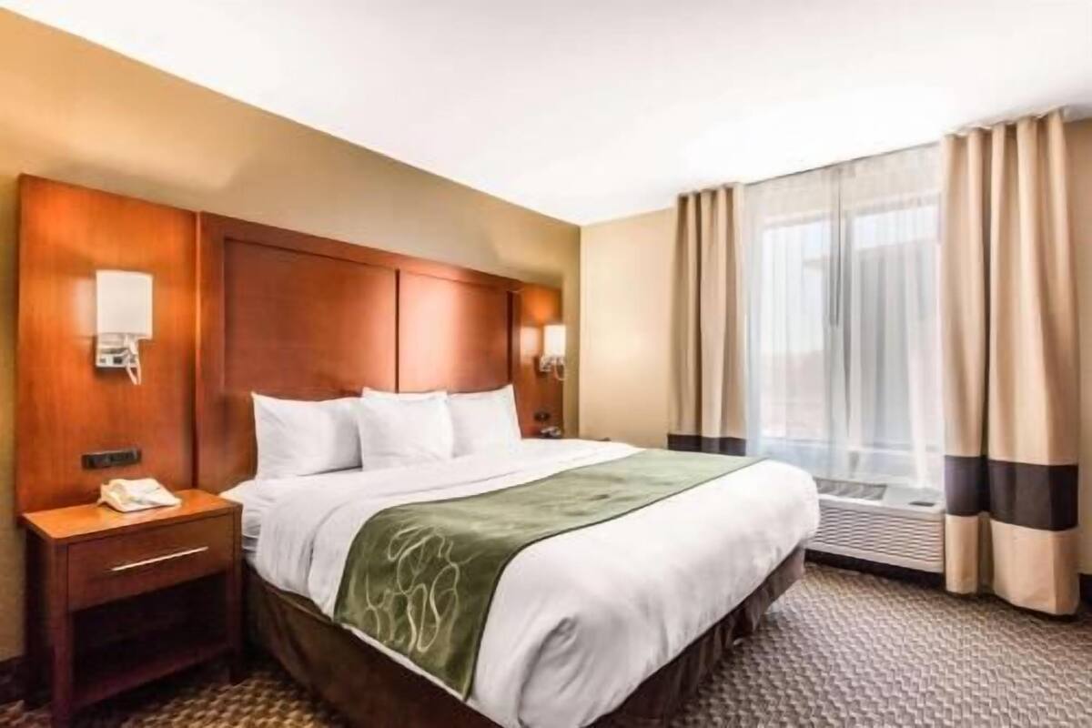 A hotel room with a bed, bedside tables, a nightstand, a window with curtains, and a wall-mounted light fixture.