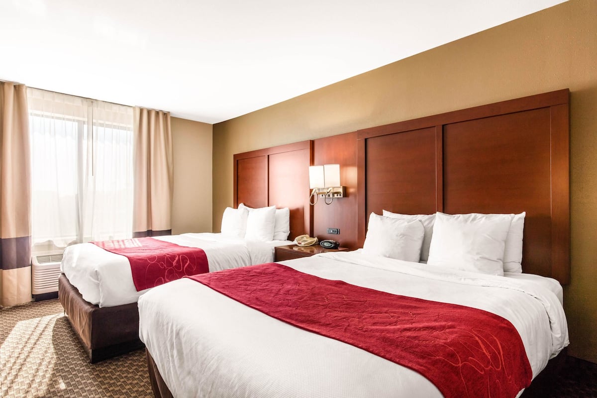 A hotel room with two beds, a wooden headboard, a bedside table with a lamp, and a window with curtains.
