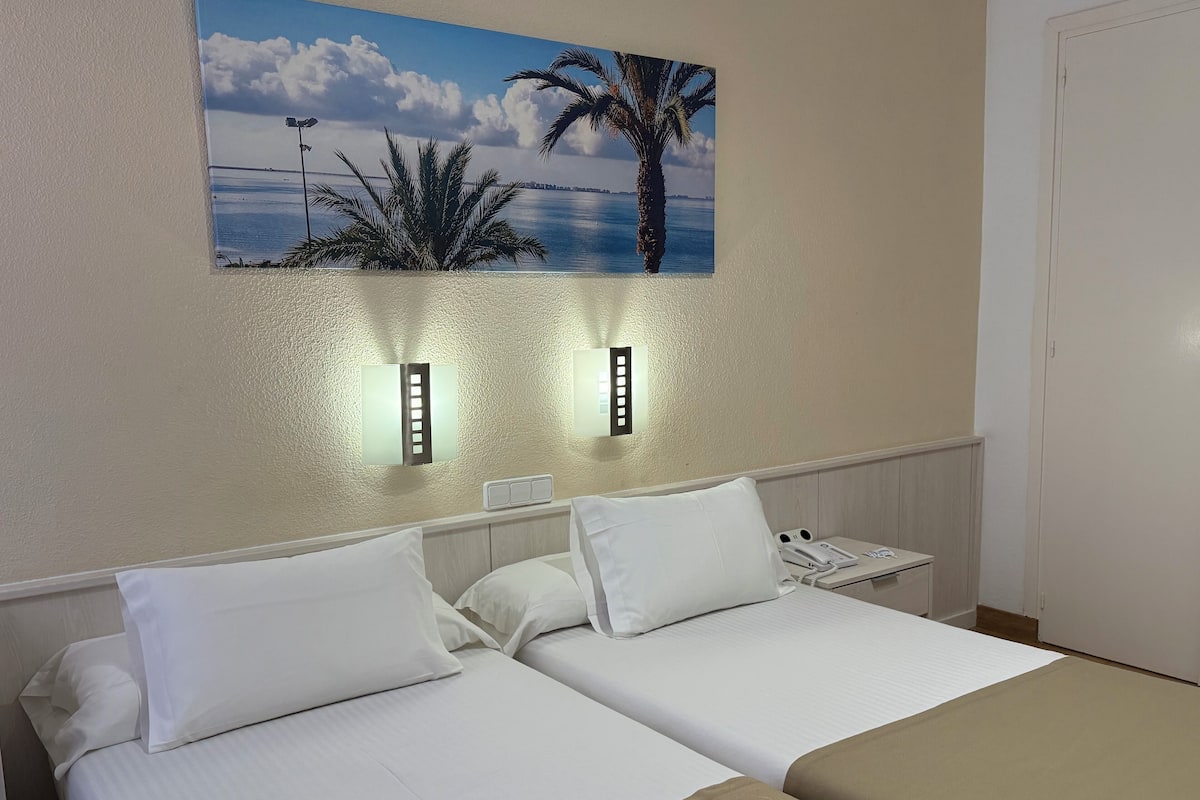 A hotel room with two beds, a painting of a beach scene, and wall-mounted lights.