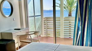 A hotel room with a bed, a desk, a chair, a small trash bin, a round mirror, and a view of the sea through large windows. - Hotel Neptuno (San Pedro del Pinatar)