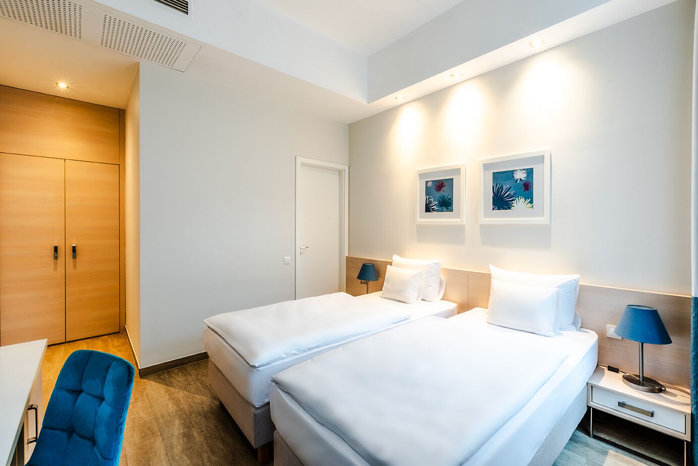 A hotel room with two beds, a desk, a chair, and artwork on the wall.