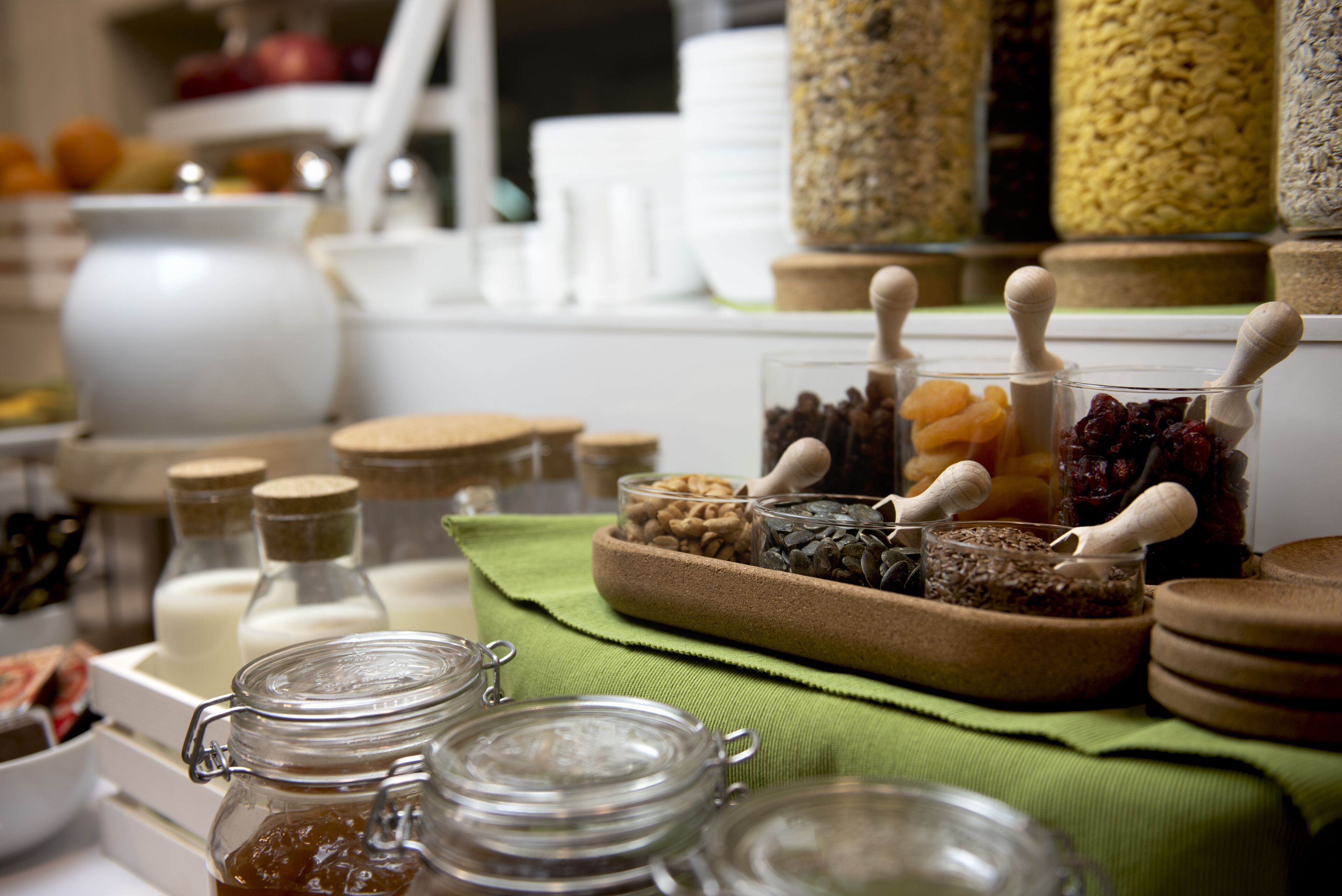 On peut voir une variété de bocaux et de récipients en verre, dotés de couvercles en bois, qui contiennent probablement des aliments ou des condiments, disposés sur une table recouverte d’une nappe verte.
