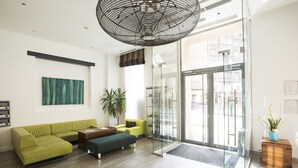 Een moderne lobby met een groene bank, een houten zitbank, een grote hanglamp en een glazen deur naar buiten.