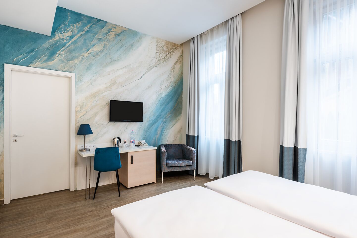 Hotellihuone, jossa on sänky, työpöytä tuolilla, televisio ja suuri abstrakti seinämaalaus.