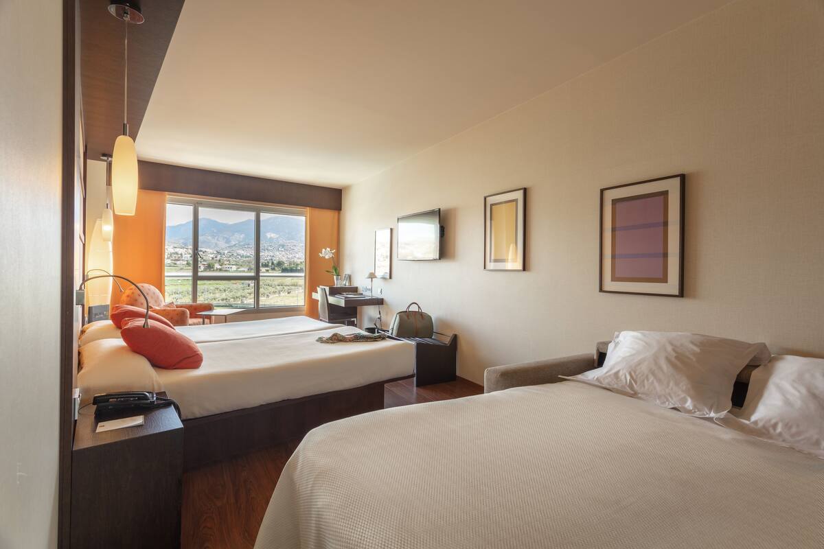 A hotel room with two beds, a desk, a chair, a window with a city view, and framed artwork on the wall.