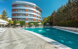 Ein Poolbereich mit Liegestühlen und Sonnenschirmen, ein modernes Gebäude mit Balkonen und ein gepflegter Heckenstreifen.