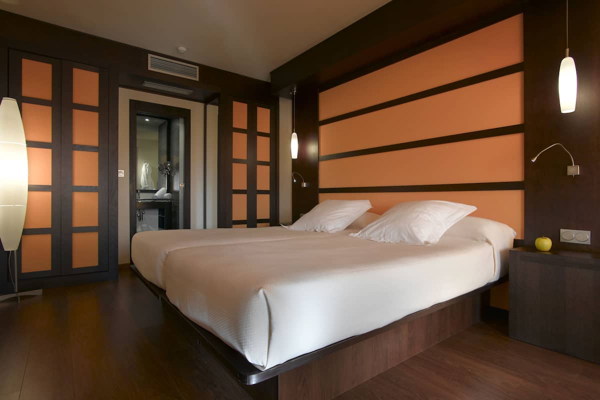 A hotel room with a large bed, wooden flooring, a nightstand with a lamp, and a wall with a geometric pattern.