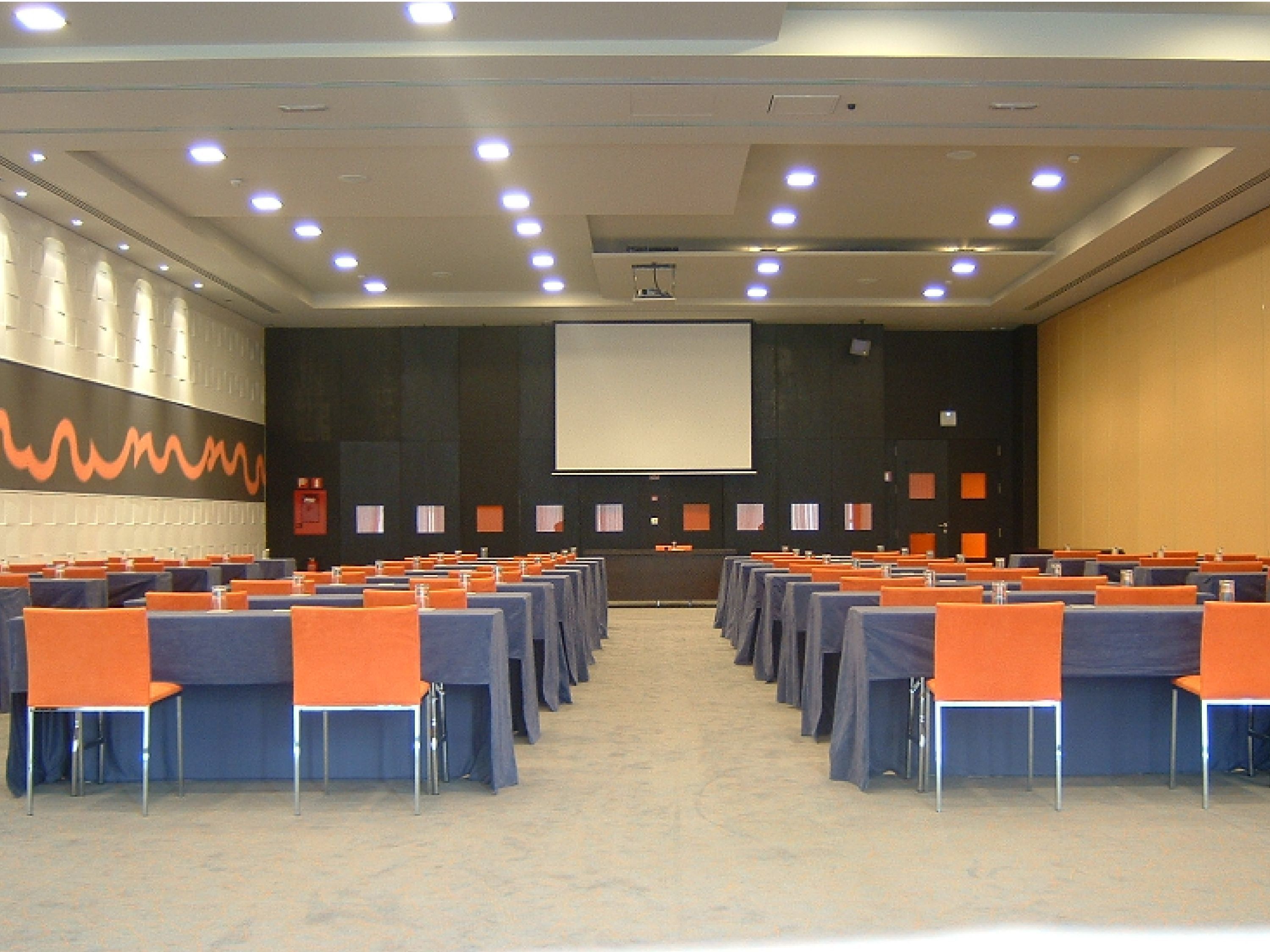 Un salón de conferencias con filas de mesas y sillas, una pantalla grande al frente y un muro con diseño.