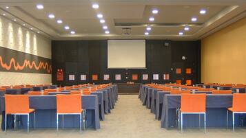 Un salón de conferencias con filas de mesas y sillas, una pantalla grande al frente y un muro con diseño.