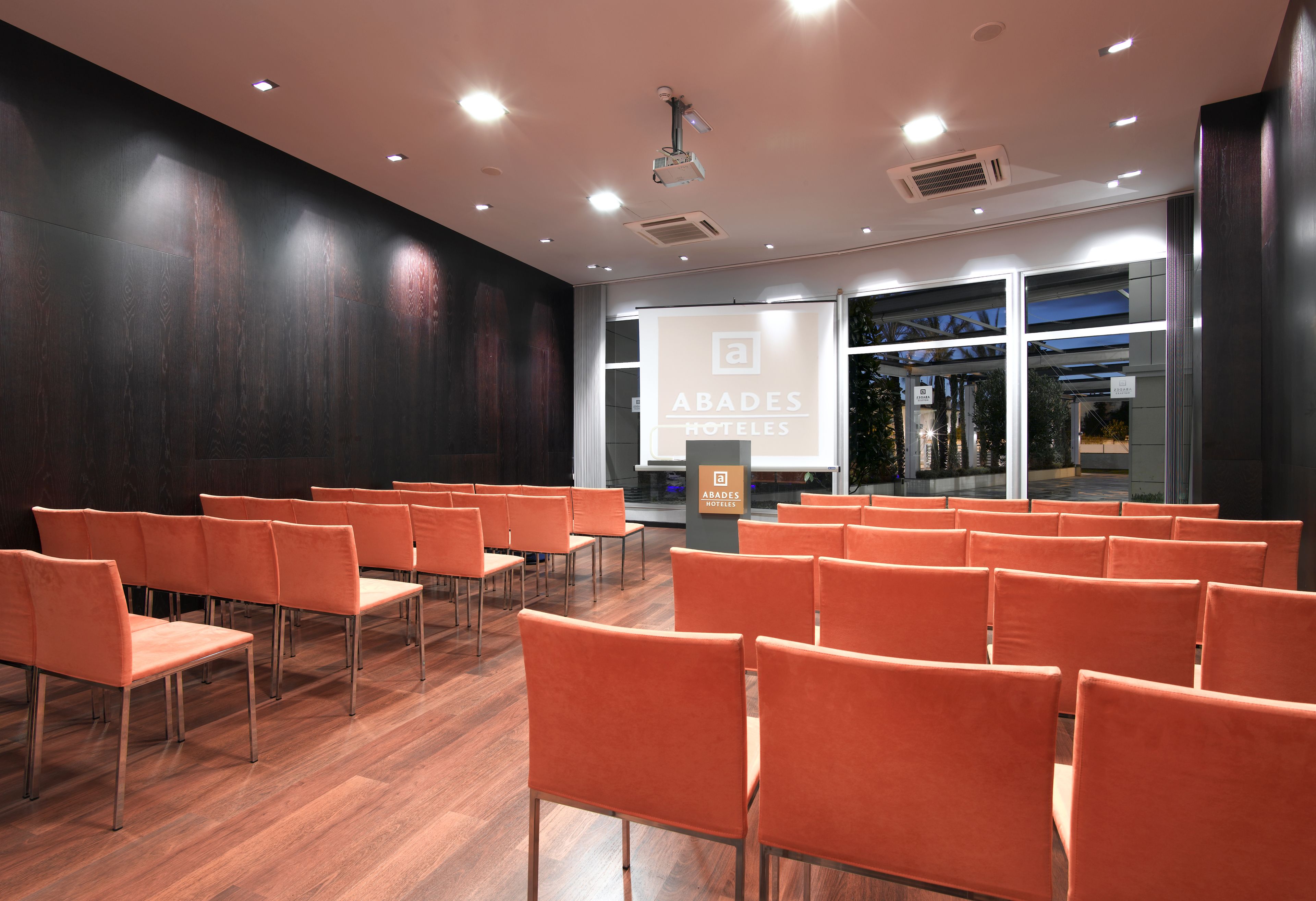 Une salle de conférence avec des rangées de chaises orange, un pupitre et un écran de projection affichant le texte « ABADES HOTELS ».