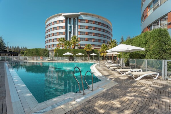 Ein Schwimmbecken mit Liegestühlen und Sonnenschirmen, umgeben von einem Gebäude mit einer geschwungenen Fassade.