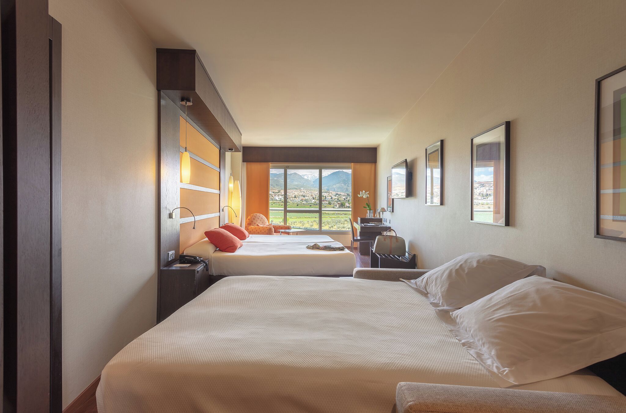 Camera d'albergo con due letti, una scrivania, una sedia e un'ampia finestra vista montagna.