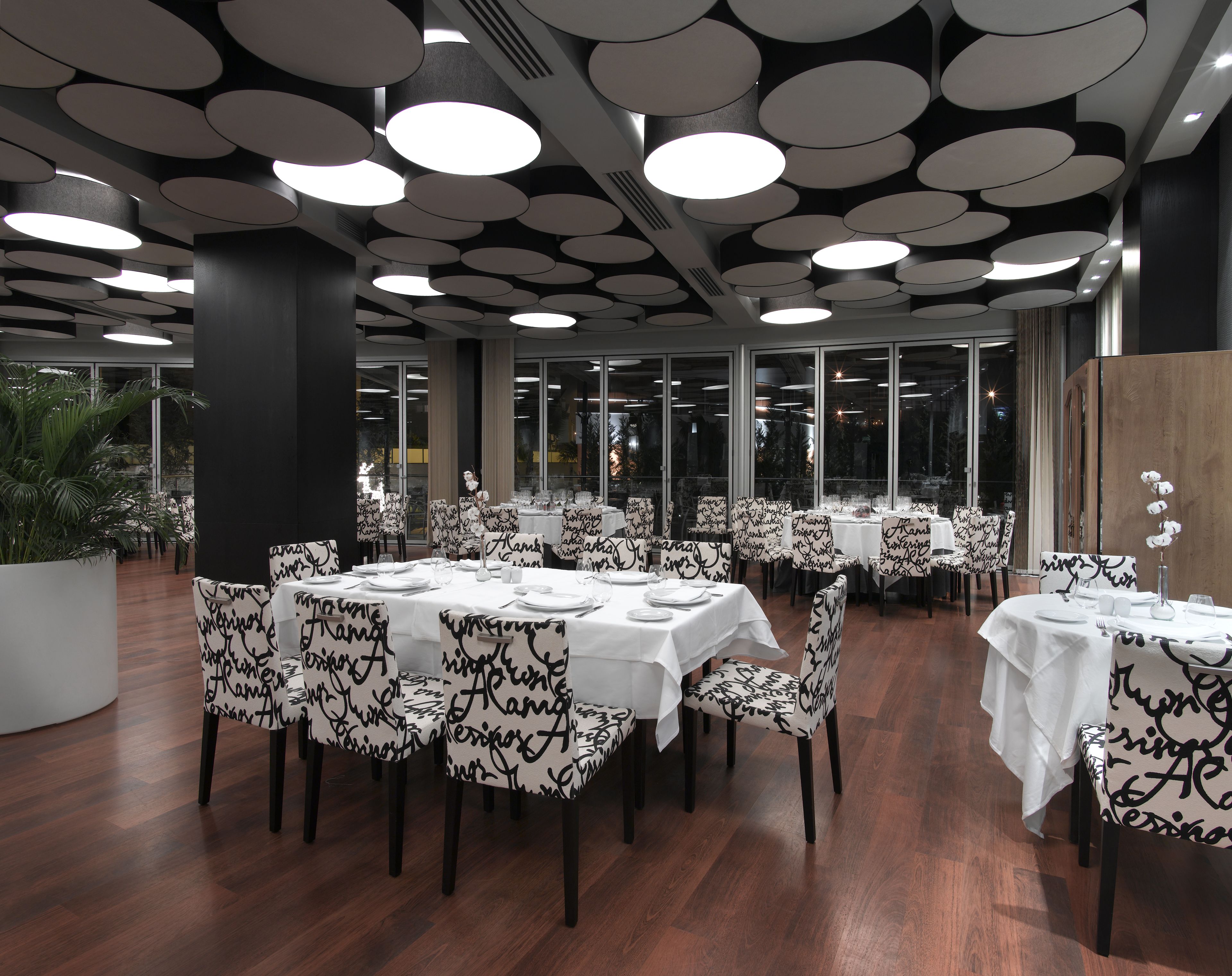 A restaurant with round tables and chairs, large windows, and a modern ceiling design.
