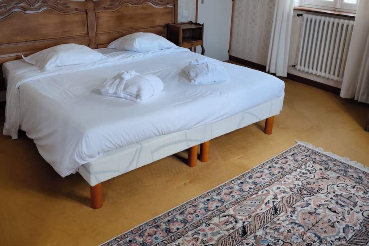 A double bed with white linens and towels, a wooden headboard, a small bedside table, a patterned rug, a framed picture on the wall, and a window with curtains.
