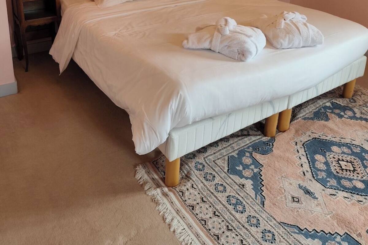 A neatly made bed with white linens and two towels on the bed. There are two bedside tables with lamps, a wooden headboard, and a patterned rug on the floor.