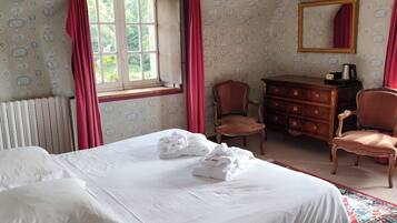 A bedroom with a large bed, a dresser, two chairs, a radiator, a window with curtains, and a chandelier.