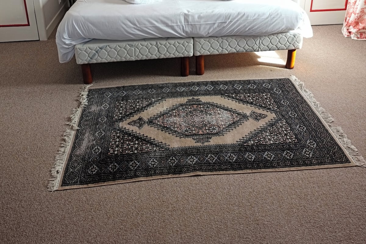 A neatly made bed with white linens, a patterned rug, and a floral wallpaper.