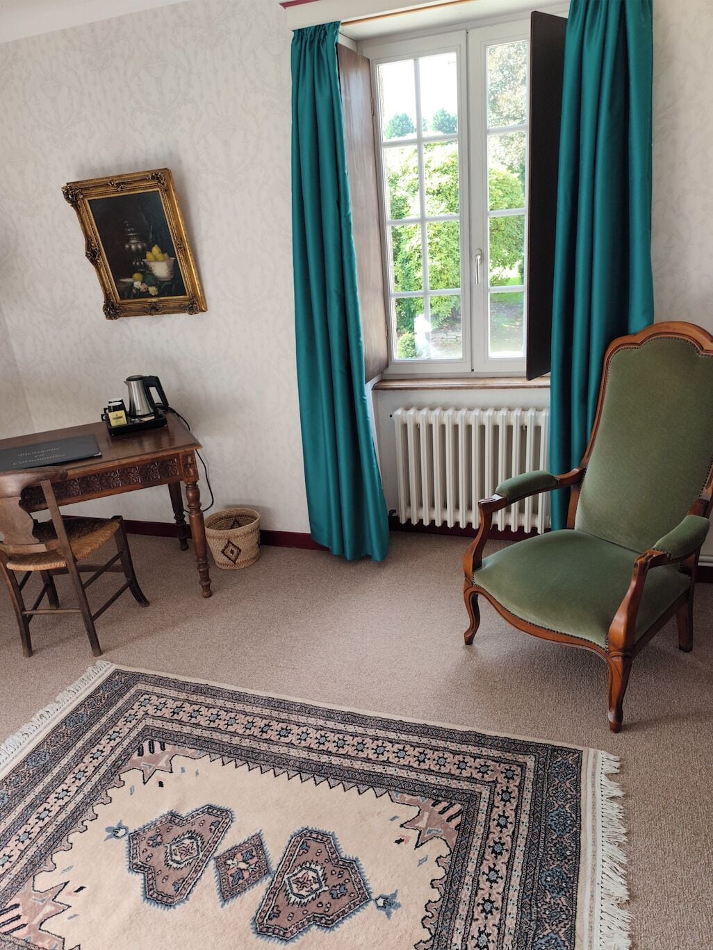 A room with a wooden desk, a green armchair, a patterned rug, a framed picture, and a window with teal curtains.
