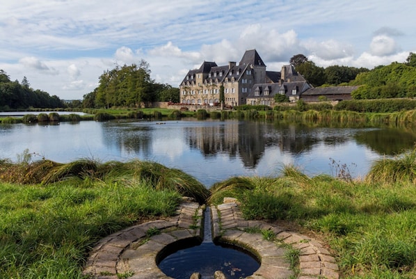 A historic building with a moat and a small bridge, surrounded by lush greenery and a calm lake.