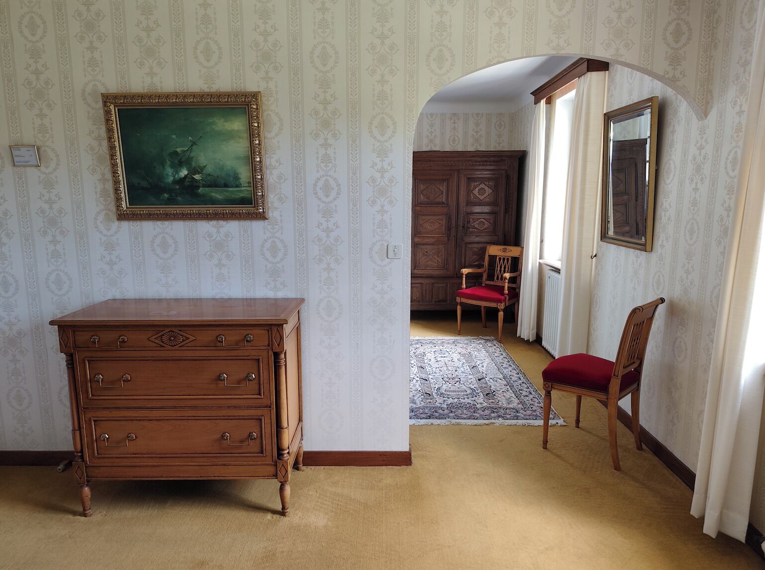 Une pièce avec une commode en bois, une chaise rouge, un tapis à motifs et un grand miroir.