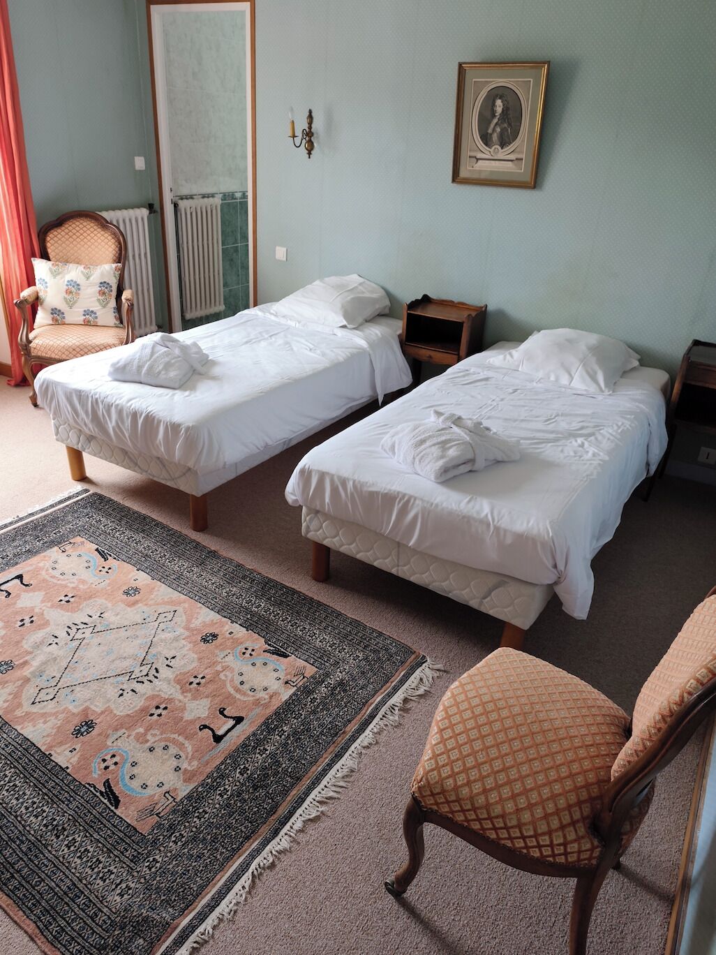A room with two single beds, a chair, a small table, a mirror, and a framed portrait on the wall.